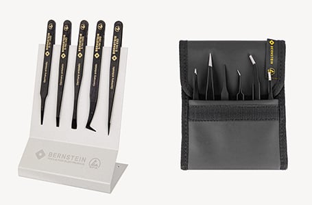 Ein fünfteiliges, schwarzes ESD-Pinzetten-Set der Marke Bernstein in einem hellgrauen Tischständer auf der linken Seite und ein weiteres Pinzetten-Set in einer geschlossenen, schwarzen Werkzeugtasche auf der rechten Seite vor weißem Hintergrund.