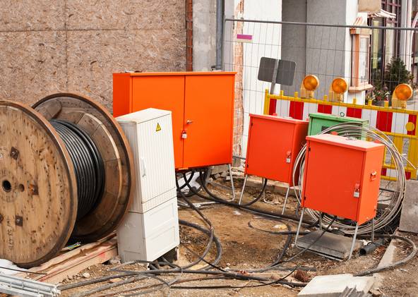 Das Bild zeigt eine Baustelle im Freien, die für Elektro- oder Versorgungsarbeiten eingerichtet ist. Auf dem Boden liegt eine große, hölzerne Kabeltrommel. Des Weiteren sind mehrere Baustromverteiler zu sehen, die in Orange lackiert sind. Diese Kästen sind auf der Baustelle verteilt und sind durch dicke schwarze Kabel, die auf dem Boden verlaufen, miteinander verbunden. Hinter den Kästen und Kabeltrommeln befindet sich eine gelb-weiß gestreifte Baustellenabsperrung mit orangefarbenen Lichtern. 