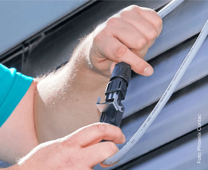  Eine Nahaufnahme zeigt zwei Hände, die ein graues Kabel an einen schwarzen Stecker vor einer Außenjalousie anschließen.