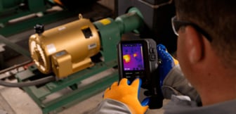 Nahaufnahme eines Technikers mit Schutzhandschuhen, der eine FLIR-Wärmebildkamera hält, um die Temperatur eines laufenden Elektromotors in einer Industrieanlage zu überprüfen.