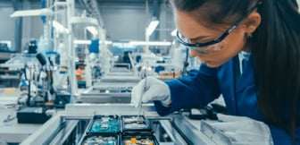 Eine Technikerin mit Schutzbrille und Handschuhen arbeitet konzentriert mit einer Pinzette an elektronischen Platinen in einer hellen, modernen Produktionshalle.
