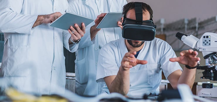 Abb. Gruppe von Ärzten, die medizinische Daten auf Tablets im Labor analysieren, im Fokus eine Person mit VR-Brille