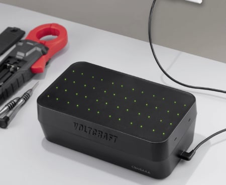 Ein schwarzes Voltcraft-Ladegerät mit zahlreichen grün leuchtenden Status-LEDs auf der Oberseite steht neben Werkzeugen wie einer Stromzange und Schraubendrehern auf einer grauen Arbeitsfläche.