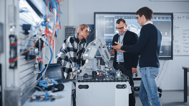 Drei Personen diskutieren über einen Roboterarm an einer Werkstation in einem modernen Labor.