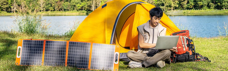 Person arbeitet am Laptop vor einem Zelt am See, mit mobilem Solarpanel für autarke Stromversorgung beim Camping.