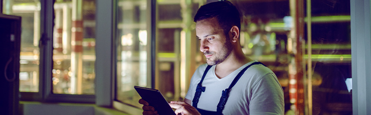 Ein Techniker in dunkler Arbeitslatzhose und weißem T-Shirt steht in einem schwach beleuchteten Technikbereich vor großen Fenstern und bedient ein Tablet. 
