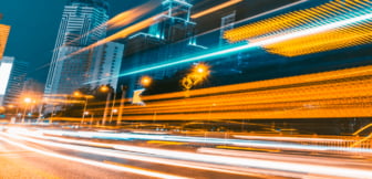 Langzeitbelichtung einer nächtlichen Stadtstraße mit dynamischen Lichtspuren vorbeifahrender Fahrzeuge vor einer Kulisse aus beleuchteten Hochhäusern.