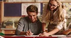 Ein junger Mann sitzt an einem Schreibtisch und schreibt konzentriert, während eine Frau über ihn gebeugt ist und ihm dabei hilft, was eine unterstützende Lernsituation darstellt.