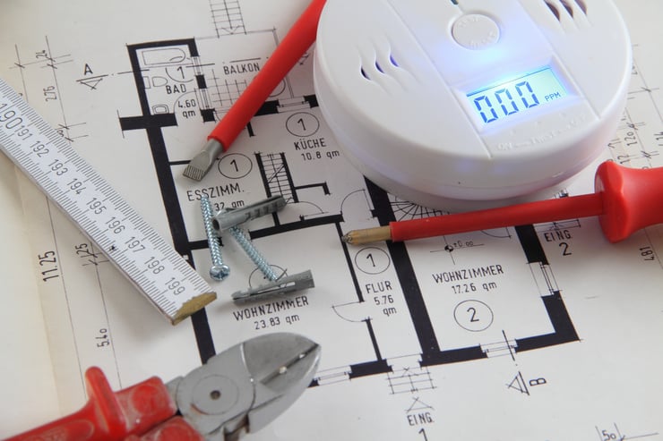 Ein Grundrissplan auf einem Tisch mit verschiedenen Schraubenziehern, einer Zange, einem Massstab und einer Brandmeldeanlage. Ebenfalls befinden sich 2 Schrauben und 2 Dübel auf dem Tisch.