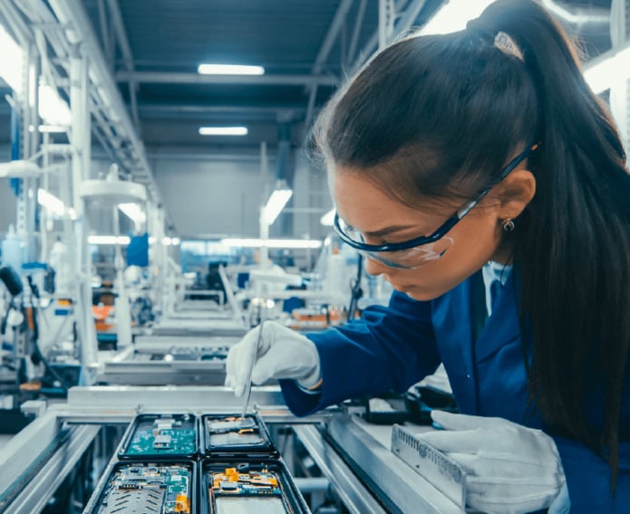 Eine Technikerin mit Schutzbrille und Handschuhen arbeitet in einer modernen Fertigungshalle konzentriert mit einer Pinzette an elektronischen Bauteilen auf einer Leiterplatte.