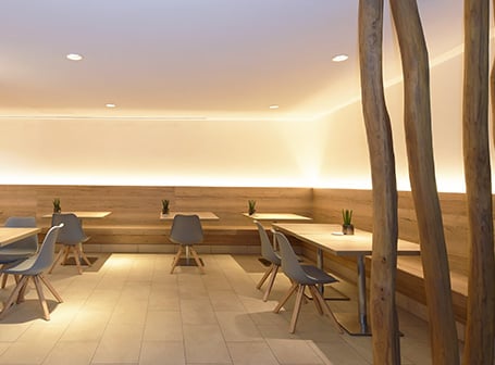 Das Innere eines Cafés oder Restaurants mit Holzbänken und Holzboden, dessen moderne, indirekte Beleuchtung durch ein durchgehendes Lichtband entsteht, das mit Aluminiumprofilen realisiert wurde.