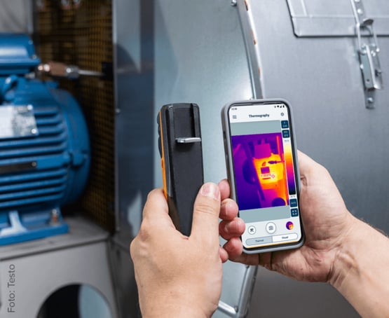 Das Bild zeigt die testo Wärmebildkamera und die dazugehörige APP auf einem Handy im Einsatz vor dem Elektromotor einer Maschine im Hintergrund.