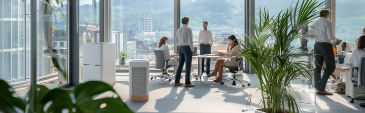 Vier Personen sitzen und stehen in einem modernen, hellen Büro mit großen Fenstern und Blick auf eine Stadtlandschaft im Hintergrund.