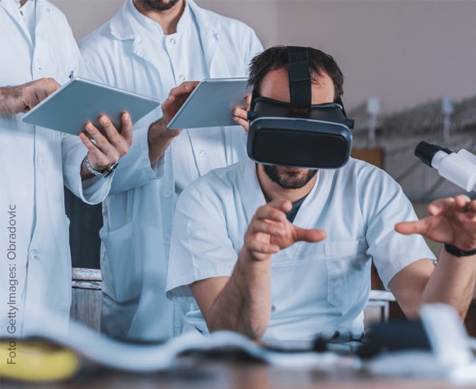 Dieses Bild zeigt Ärzte in weißen Laborkitteln, die mit Virtual-Reality-Technologie arbeiten. Eine Person trägt ein VR-Headset und ist in eine Simulation vertieft, während zwei weitere Personen mit Tablets daneben stehen.