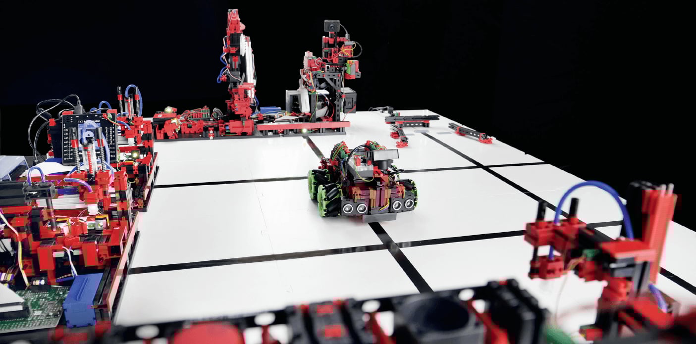 Ein kleiner, aus roten und schwarzen Bauteilen konstruierter Roboter mit grünen Rädern navigiert auf einer weißen Fläche mit schwarzen Linien zwischen verschiedenen stationären modularen Technik-Aufbauten.