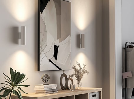  Eine helle, moderne Flur- oder Wohnzimmereinrichtung mit einer Konsole, auf der Bücher und Dekoration stehen, einem langen, abstrakten Bild an der Wand, das von zwei up-and-down Wandleuchten beleuchtet wird, die warmes Licht nach oben und unten werfen.