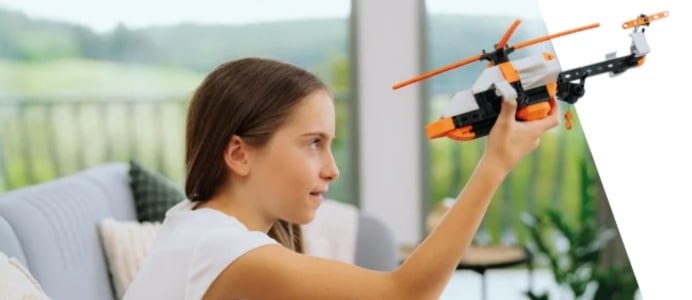 Ein Mädchen mit langen braunen Haaren sitzt lächelnd auf einem Sofa und hält ein weißes, orangefarbenes und schwarzes Hubschraubermodell aus fischertechnik-Bauteilen in die Höhe.