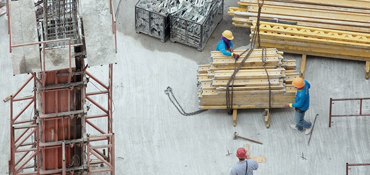 Arbeiter mit Schutzhelmen bewegen auf einer Baustelle mit einem Kran eine gestapelte Last aus Baugerüstteilen.