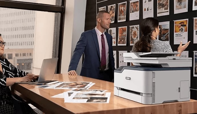 Drei Personen arbeiten in einem Büro an einem Holztisch mit einem Laptop und Ausdrucken, während im Vordergrund rechts ein grauer Xerox Multifunktionsdrucker steht und an der Wand im Hintergrund mehrere Ausdrucke hängen.