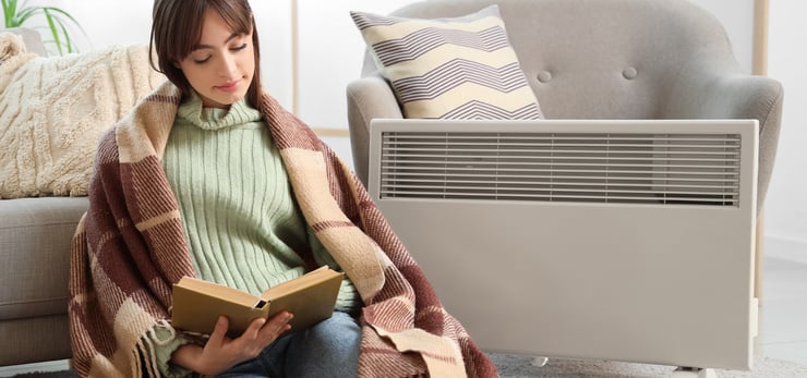 Frau liest entspannt ein Buch neben einem elektrischen Heizgerät in einem gemütlich eingerichteten Wohnzimmer.