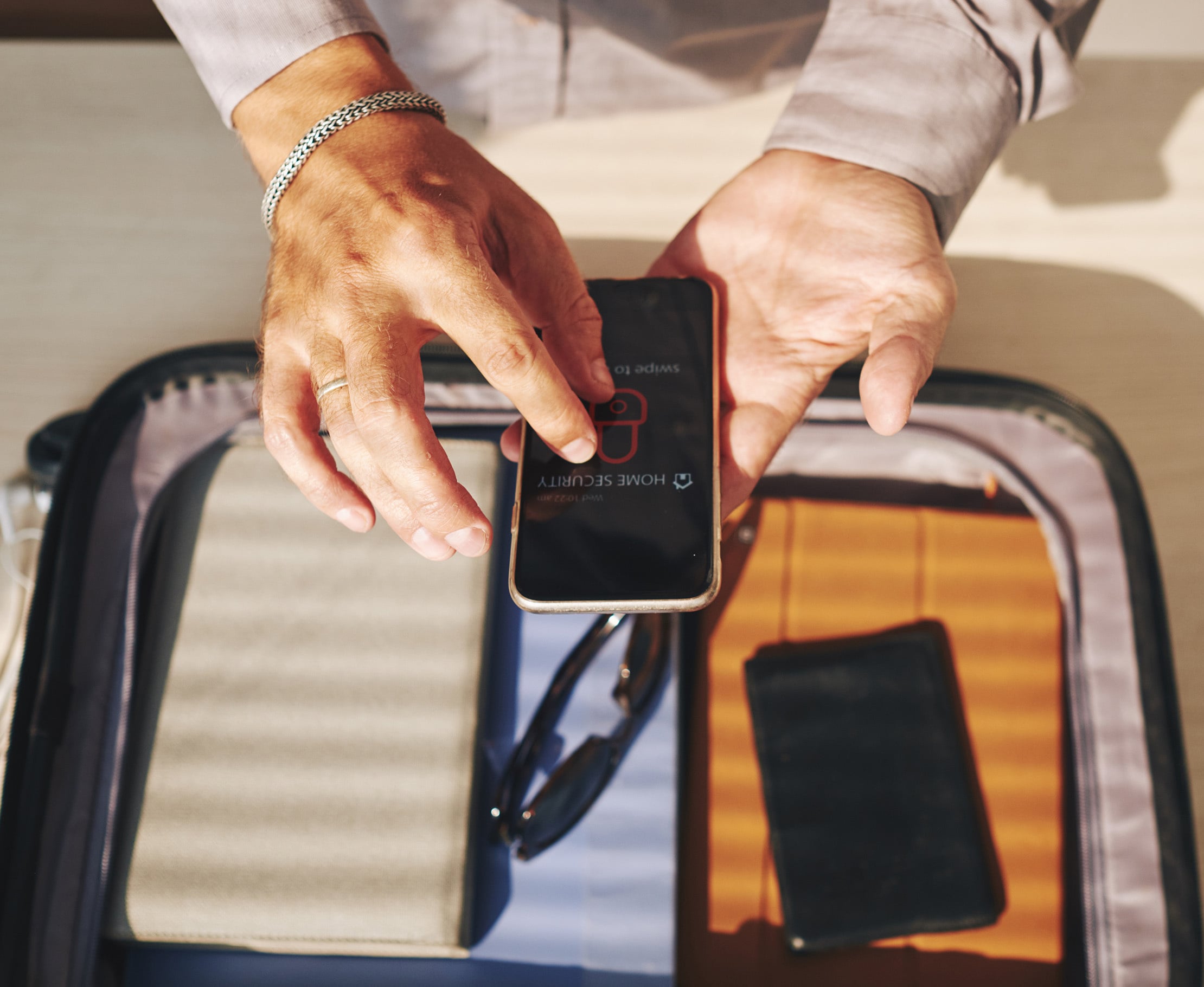 Werbegrafik mit einer Person, die eine Sicherheits-App auf dem Smartphone nutzt, und dem Text ‚Sicherheit für Ihren Urlaub‘ für den Schutz persönlicher Wertsachen auf Reisen.