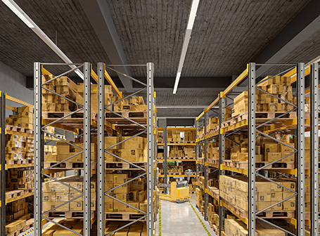  Das Bild zeigt eine breite Gasse in einem großen Lagerhaus mit hohen, silbernen Regalen voller verpackter Kartons auf Holzpaletten, die von zwei langen, durchgehenden, hellen Lichtbandsystemen an der Decke gleichmäßig beleuchtet werden.