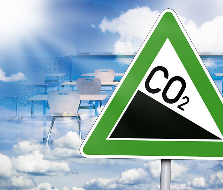 Ein grünes CO₂-Warnschild steht vor einem leeren Klassenzimmer mit überlagerten Wolken und Sonnenstrahlen.