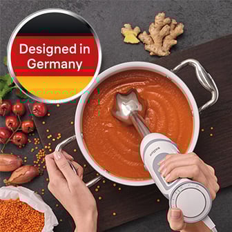 Das Bild zeigt eine Nahaufnahme von zwei Händen, die einen weißen und grauen Stabmixer in einem weißen Topf mit orangefarbener Suppe halten, umgeben von Zutaten wie roten Tomaten, Ingwer, Schalotten und roten Linsen auf einer dunklen Oberfläche, mit einem runden Aufkleber oben links, der die deutsche Flagge und den Text "Designed in Germany" zeigt.