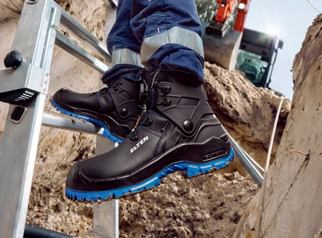 Eine Person in Arbeitskleidung steht auf einer Aluminiumleiter vor einer Baustelle und trägt schwarze ELTEN-Sicherheitsstiefel mit blauer Profilsohle und integrierter BIOMEX-Knöchelstütze.