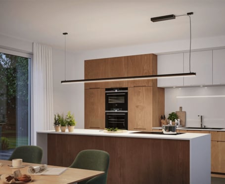 Anwendungsbeispiel 2: Das Bild zeigt das Lichtkanalsystem als schwebendes Element von der Decke abgehängt montiert in einer offenenen, modernen Küche mit geschmackvollen Möbeln in warmen Holz-Tönen. Der Kochtresen der Küche ist mit einer weissen Arbeitsplatte ausgestattet. Im Vordergrund steht ein gedeckter Esstisch mit gepolsterten Stühlen.