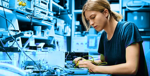 Eine junge Frau arbeitet konzentriert an elektronischen Geräten in einem Labor, umgeben von Technik. Das Bild ist in Blautönen gehalten.