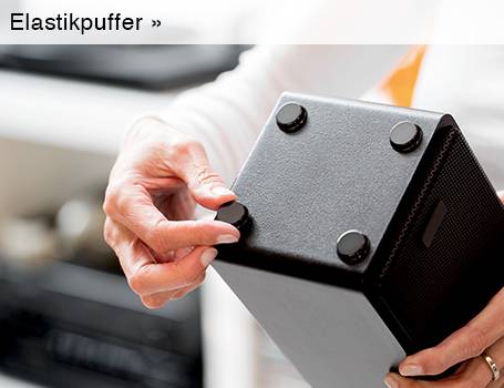 Eine Person hält einen schwarzen, quaderförmigen Gegenstand in den Händen und befestigt mit Daumen und Zeigefinger einen kleinen, runden schwarzen Elastikpuffer an einer der vier Ecken der Unterseite; der Text "Elastikpuffer »" im oberen linken Bereich des Bildes weist auf eine Weiterleitung hin.