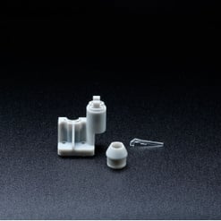 Das Bild zeigt drei kleine, graue Kunststoffteile unterschiedlicher Form auf dunklem, strukturiertem Hintergrund, darunter ein h-förmiges und ein zylindrisches Teil.