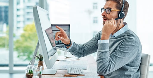 ein Mann mit Headset und Brillle sitzt am Arbeitsplatz und tippt  mit der Hand auf seinen Touchscreen