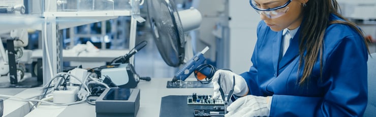 Frau in blauer Arbeitskleidung und weissen Handschuhen arbeitet an einer Elektronikplatine in einer modernen Fabrikumgebung