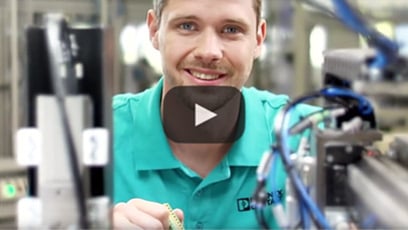 Un homme en chemise turquoise sourit devant une machine avec un bouton de lecture superposé devant lui.