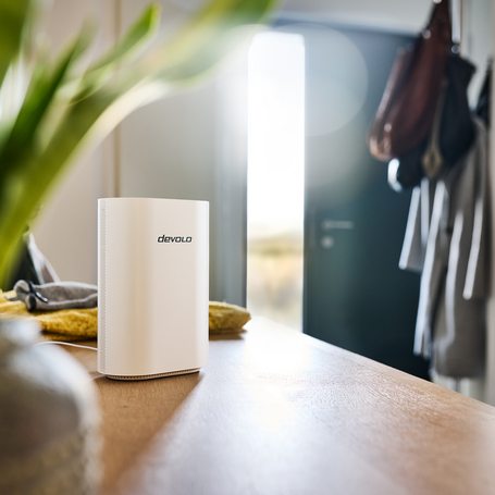 Ein weißer devolo WLAN-Router steht auf einer Holzoberfläche im Flur, mit einer hellen Haustür im unscharfen Hintergrund.