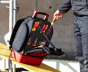 Das Bild zeigt eine Person, die einen Werkzeugtasche auf einer Baustelle hält. Die Tasche enthält verschiedene Elektrowerkzeuge, was auf handwerkliche für Elektriker. Solche Taschen sind darauf ausgelegt, Werkzeuge sicher und organisiert zu transportieren. 