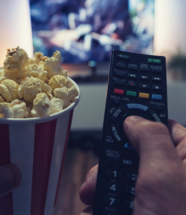 Nahaufnahme einer Hand mit Fernbedienung und einer Popcornschale vor einem laufenden Fernseher.