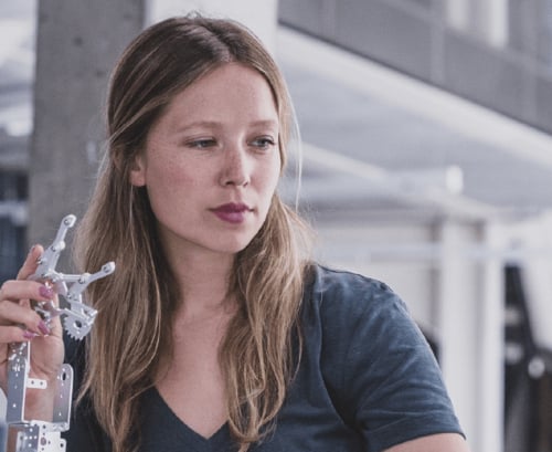 Eine junge Frau mit langen blonden Haaren blickt konzentriert auf einen filigranen, metallischen Roboterarm, den sie in ihrer Hand hält.