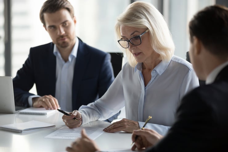 Drei Personen sitzen an einem Tisch in einem Meeting, eine ältere Frau liest ein Dokument, während die beiden jüngeren Personen zuhören.