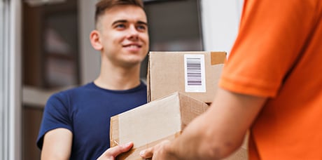  Das Bild zeigt eine Übergabe-Szene: Ein lächelnder junger Mann in einem dunkelblauen T-Shirt (links, scharf) nimmt Pakete von einer Person (rechts, unscharf) mit orangefarbenem Oberteil entgegen, die die Pakete reicht.