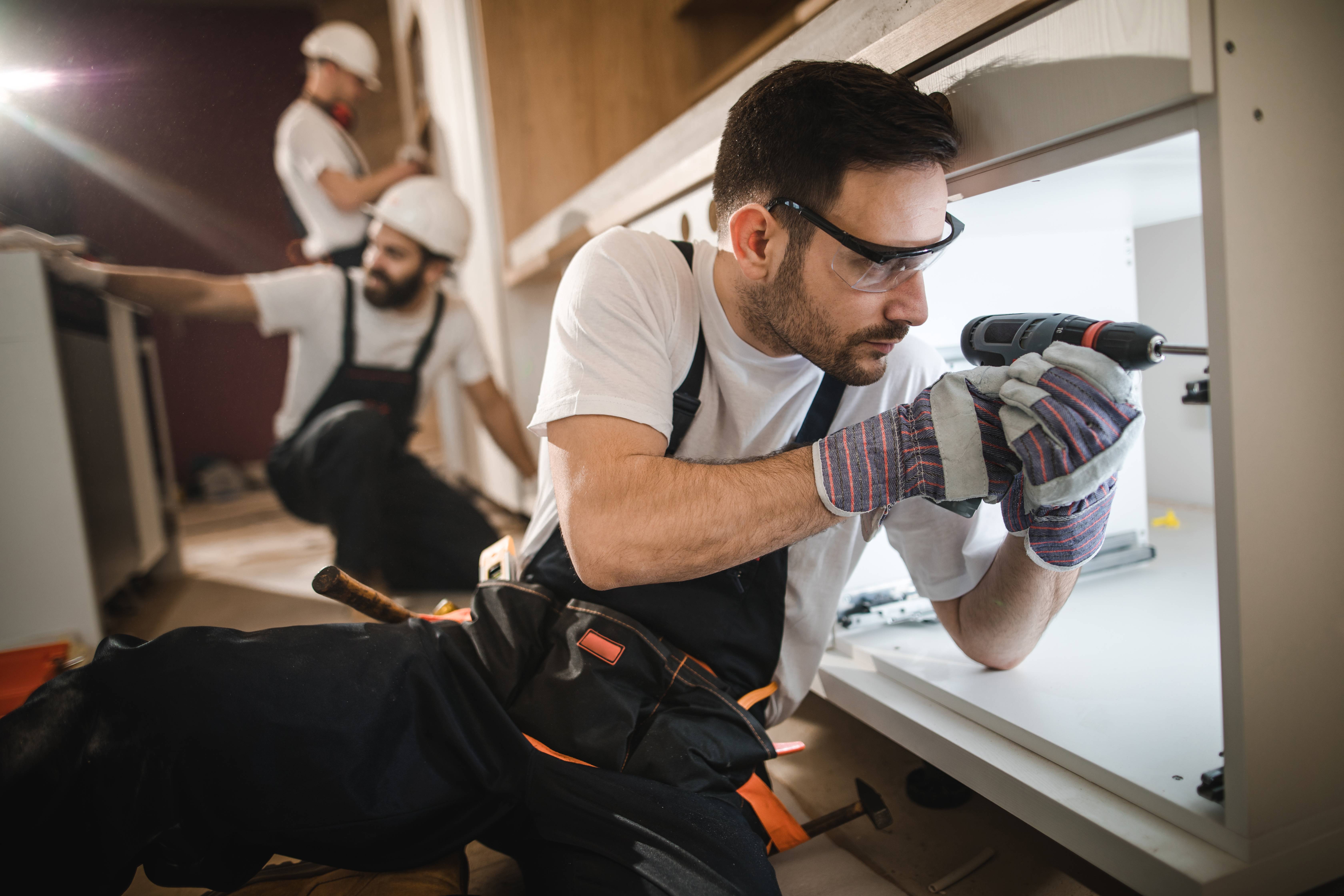 Handwerker repariert eine Küchenschublade, während im Hintergrund weitere Handwerker arbeiten.