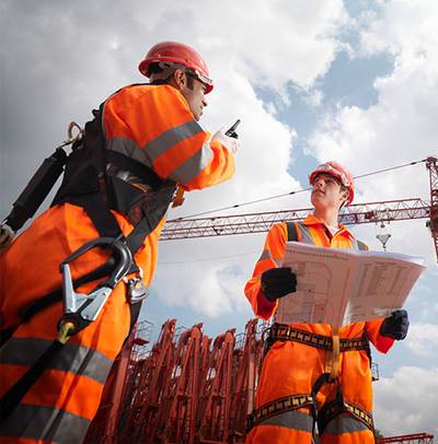 Deux ouvriers de construction en tenue de protection orange et casques discutent sur un chantier avec des grues en arrière-plan, l’un tenant des plans de construction.
