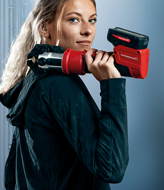 Eine Handwerkerin blickt in die Kamera und trägt eine rote Akku-Rohrpressmaschine auf ihrer Schulter.