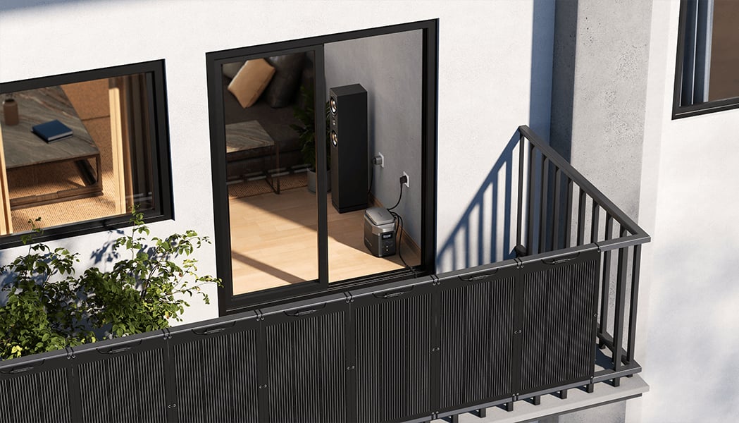 Balkon mit schwarzem Geländer und Sichtschutz. Eine Powerstation steht neben einer offenen Schiebetür, die in ein Wohnzimmer mit Sofa und Lautsprecher führt. Draußen sind Pflanzen sichtbar.