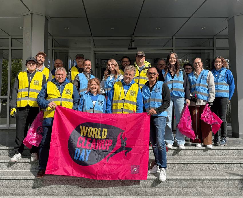 Eine Gruppe von Conrad Electronic Mitarbeitern hält vor einem Gebäude ein pinkfarbenes Banner mit der Aufschrift „World Cleanup Day“.