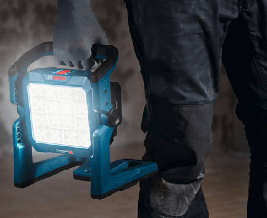 Eine Person trägt eine hell leuchtende, blau-schwarze Akku-Bauleuchte GLI 18 V-5000 von Bosch Professional am Tragegriff.