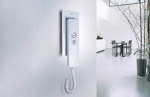 Die Kamera ist auf die Innenstation einer Türsprechanlage in einer Wohnung gerichtet. Die Station ist weiß und verfügt über einen kabelgebundenen Hörer. Im Hintergrund befinden sich ein Regal, ein Esstisch und Pflanzen.