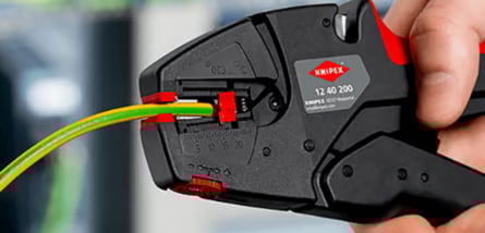  Eine Nahaufnahme zeigt eine Hand, die mit einem schwarzen und roten Werkzeug der Marke KNIPEX ein grün-gelbes Stromkabel abisoliert.